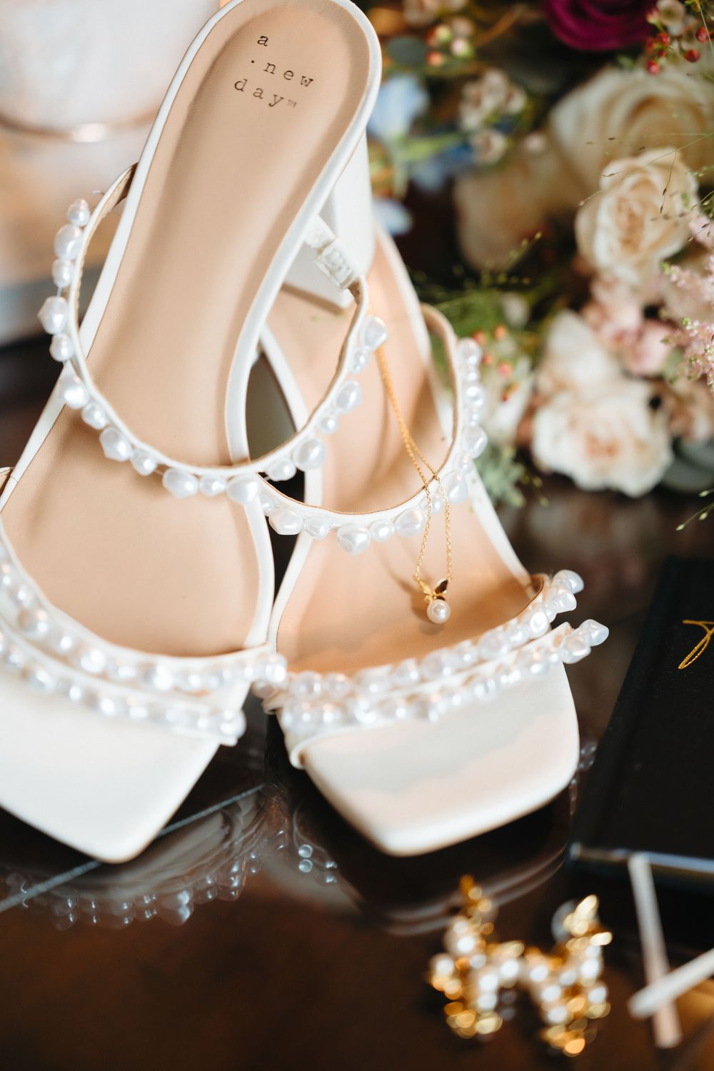 White pearl-embellished bridal heels with gold necklace on glass table near floral bouquet.
