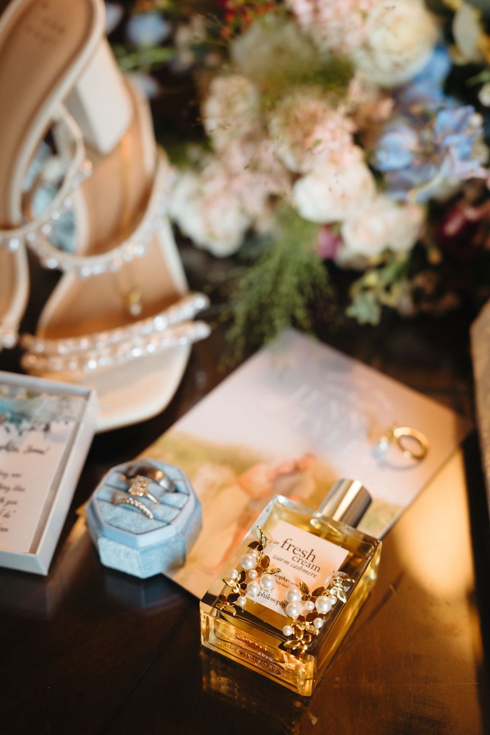 Bridal flat lay with Fresh perfume, diamond rings in blue velvet box, jeweled heels, gold band, and floral bouquet on dark wood.