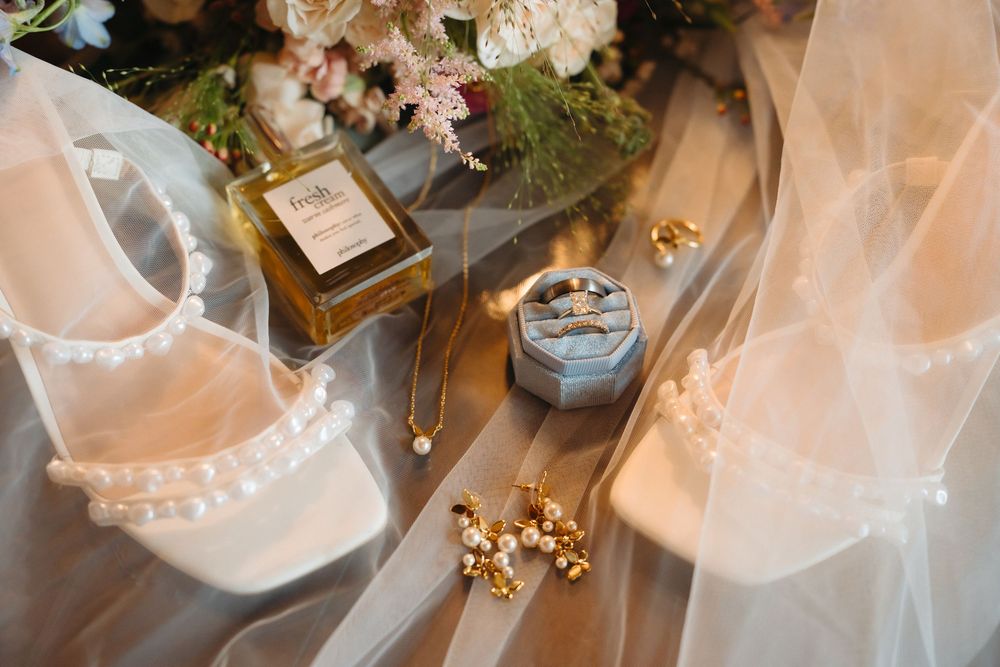 Elegant bridal flat lay featuring pearl heels, perfume, ring box, gold jewelry, and floral bouquet on soft tulle.