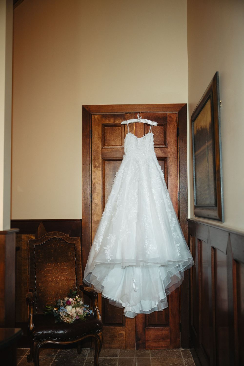 White lace wedding gown hanging on wooden door with floral bouquet on antique chair nearby.
