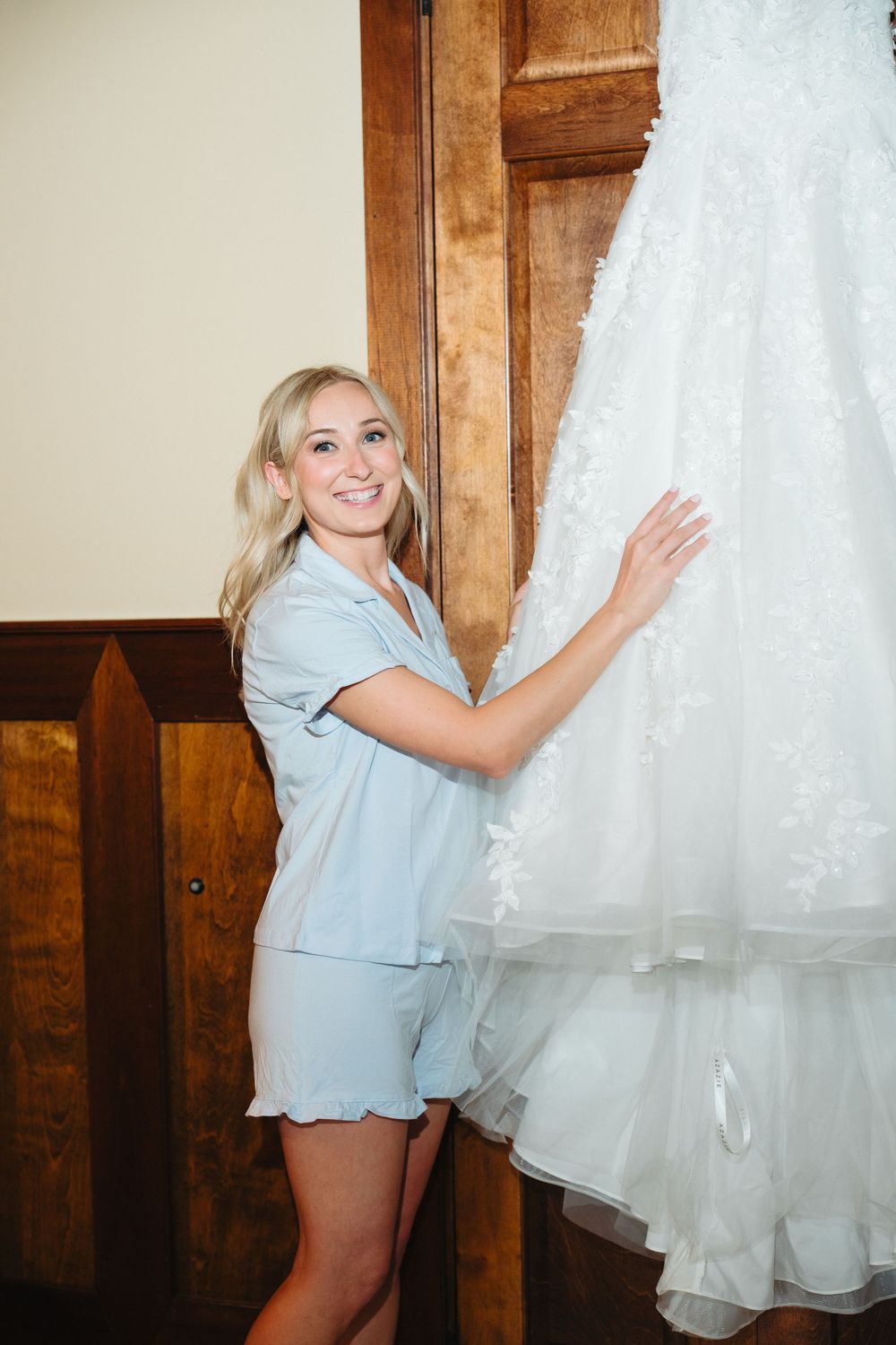 Smiling blonde bridesmaid in blue pajamas touching a white lace wedding dress hanging on a wooden door.