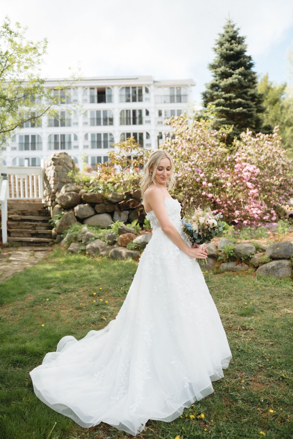 Bride in white ball gown holding bouquet in garden with blooming flowers and elegant building backdrop.
