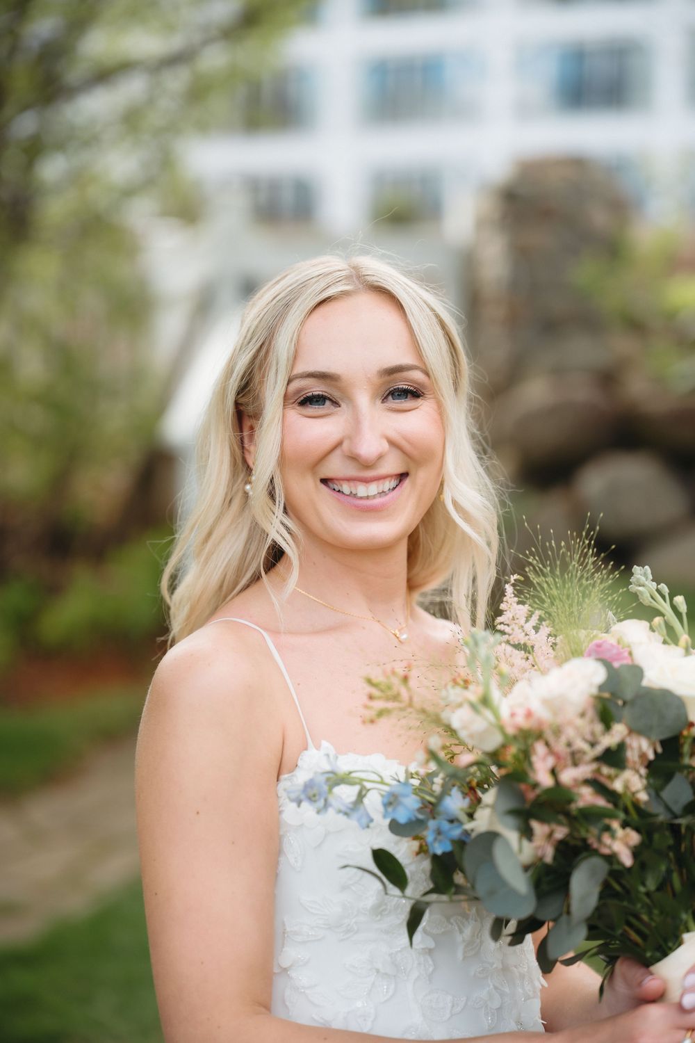 Smiling blonde bride in white dress holding a bouquet of pink, blue, and white flowers outdoors.
