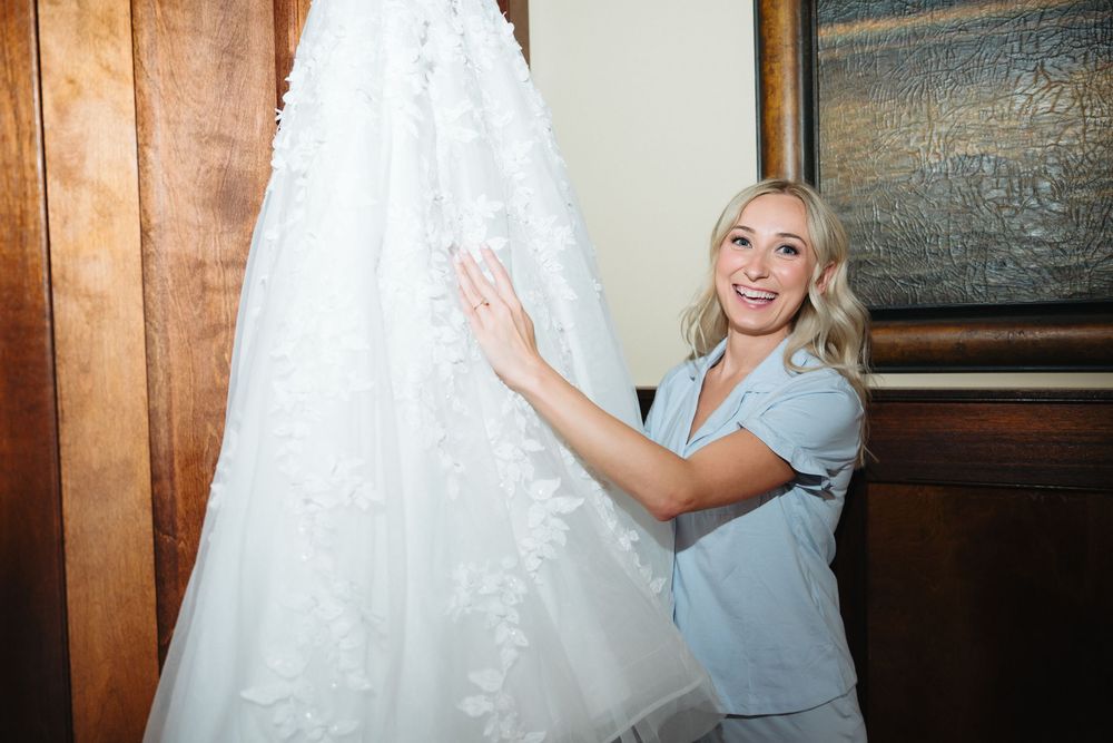 Smiling blonde woman in blue top touching elegant white lace wedding dress hanging on wooden door.