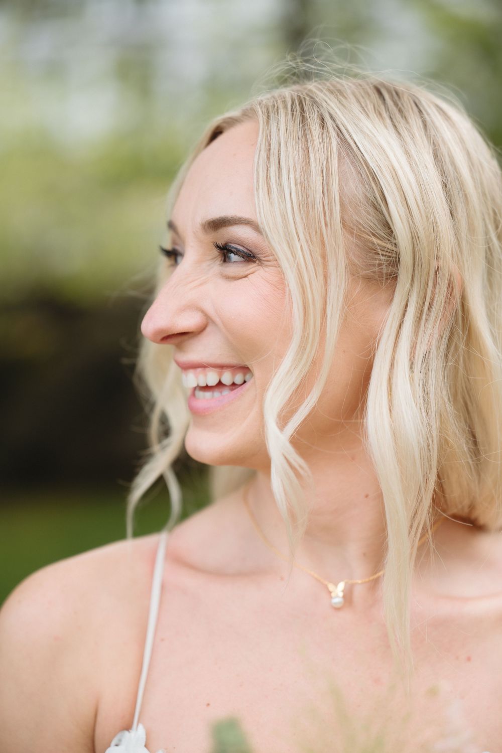 Smiling blonde bride laughing outdoors wearing a delicate gold necklace on her wedding day.