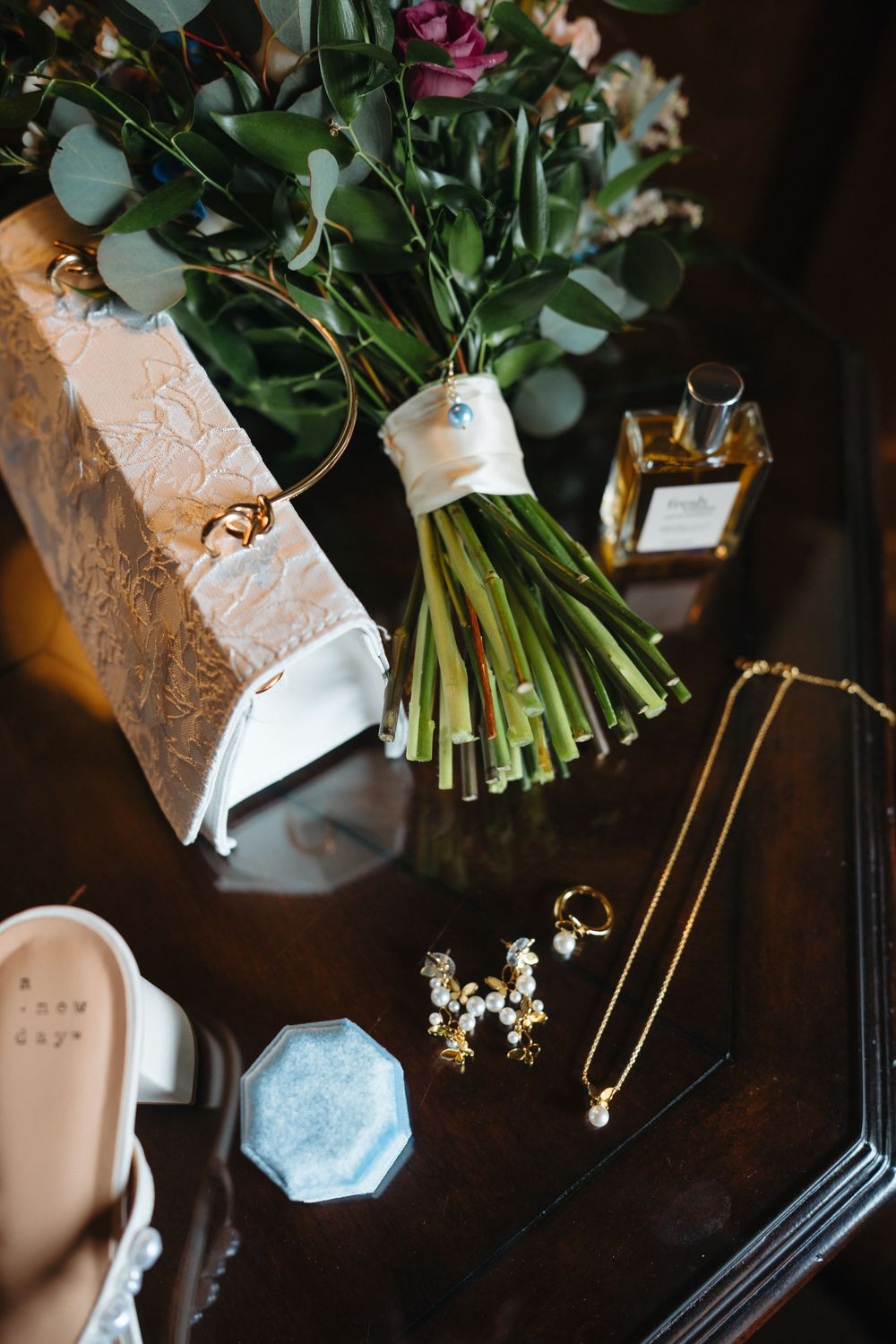 Bridal flat lay featuring white heels, gold jewelry, perfume bottle, floral bouquet, and a blue ring dish on a dark wood surface.