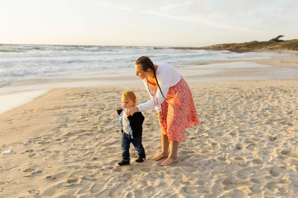 A mother helps her toddler walk on a sandy beach at golden hour with ocean waves in the background.