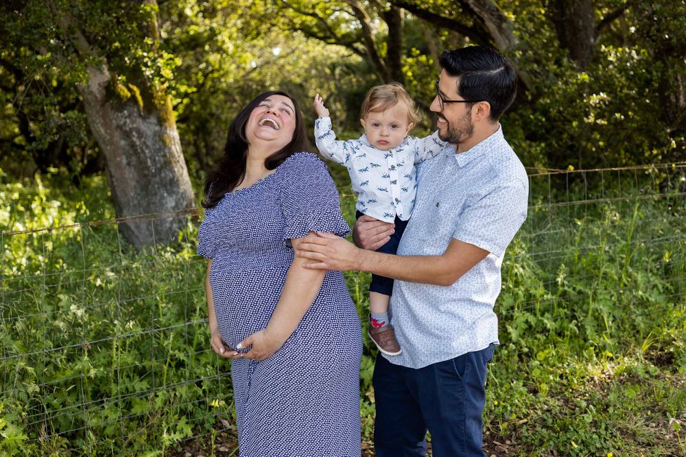 Pregnant woman in blue dress laughs with husband holding toddler outdoors in lush green park setting.