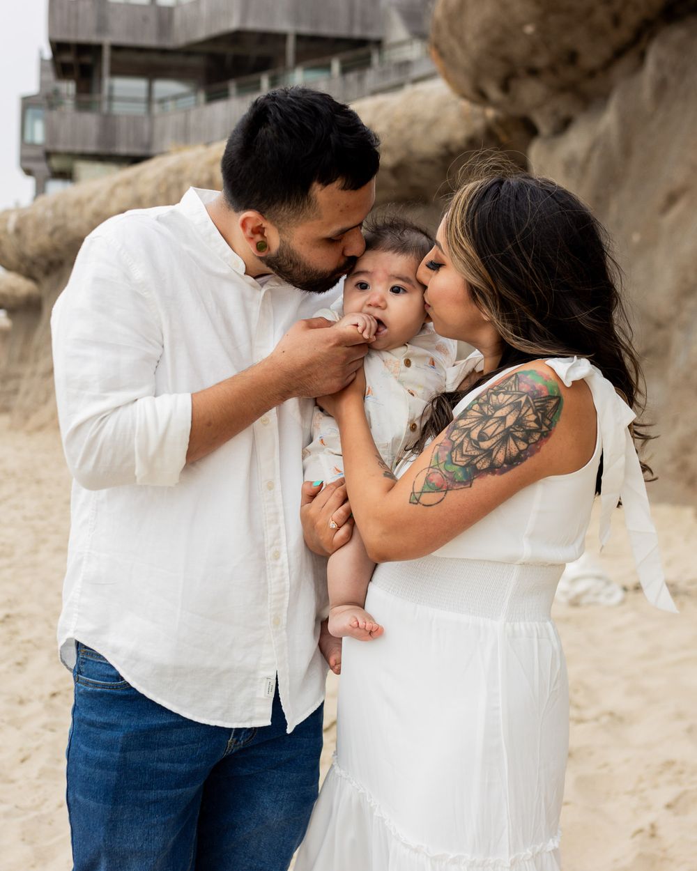 A couple in white outfits kisses their baby on a sandy beach with rocky cliffs in the background.