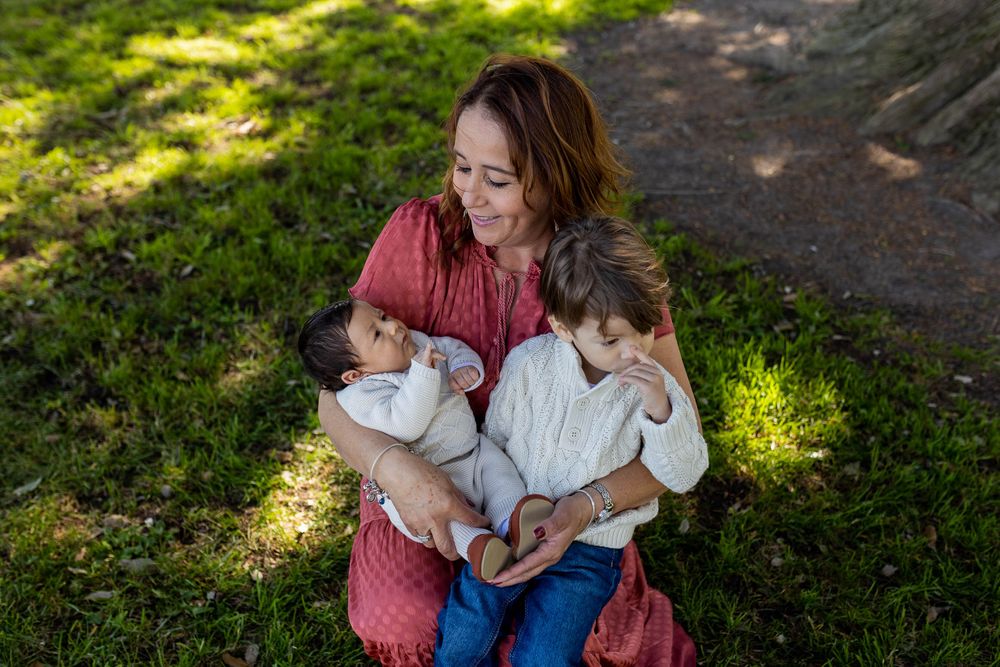 A woman sits on grass in a park, lovingly holding a newborn and a toddler in her arms on a sunny day.