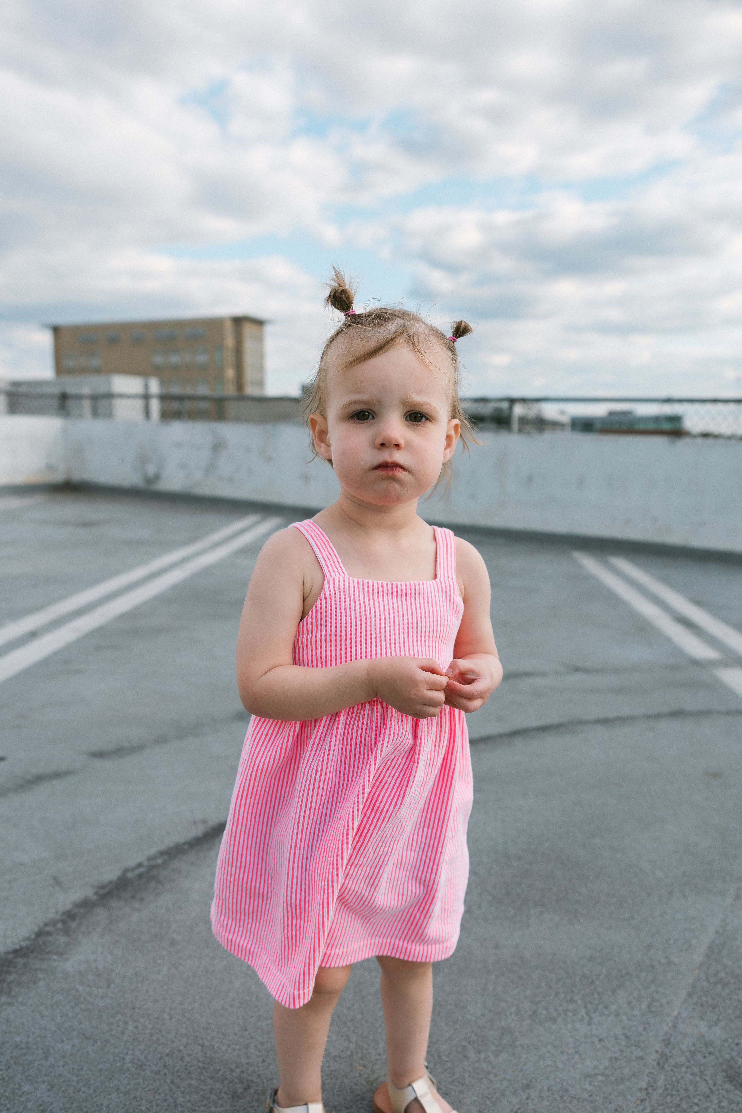 A toddler girl in a pink striped sundress stands in a parking lot, her hair in two small buns, looking at the camera.