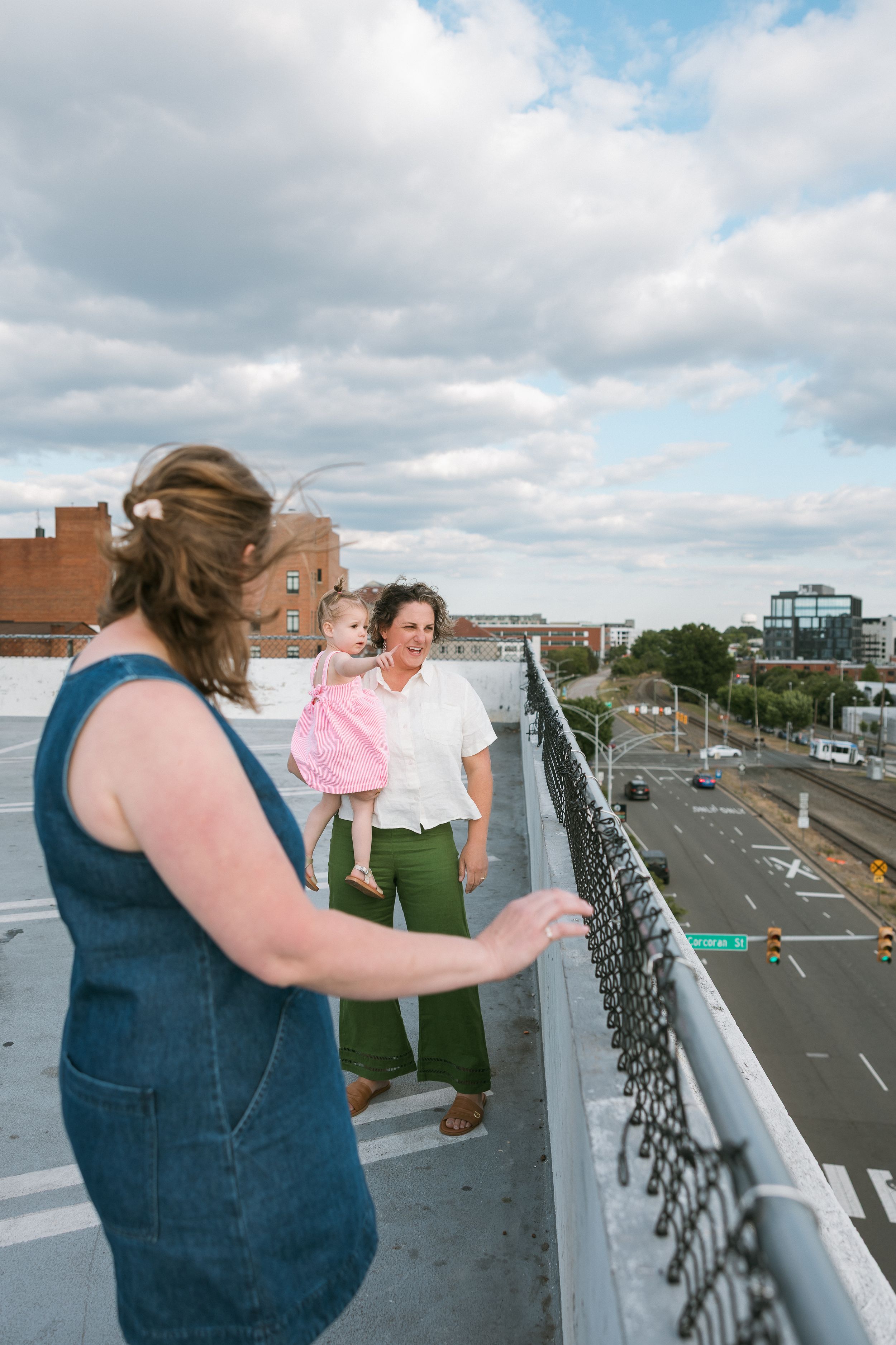 Two women and a toddler on a rooftop overlooking a city street on a cloudy day.