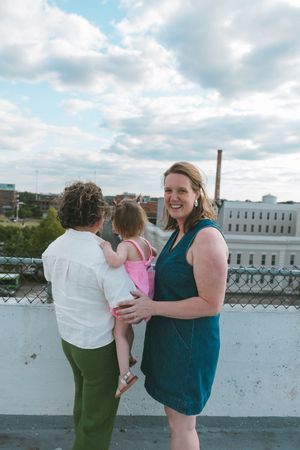 Two women and a toddler girl pose on a rooftop terrace with a cloudy sky and urban buildings behind them.