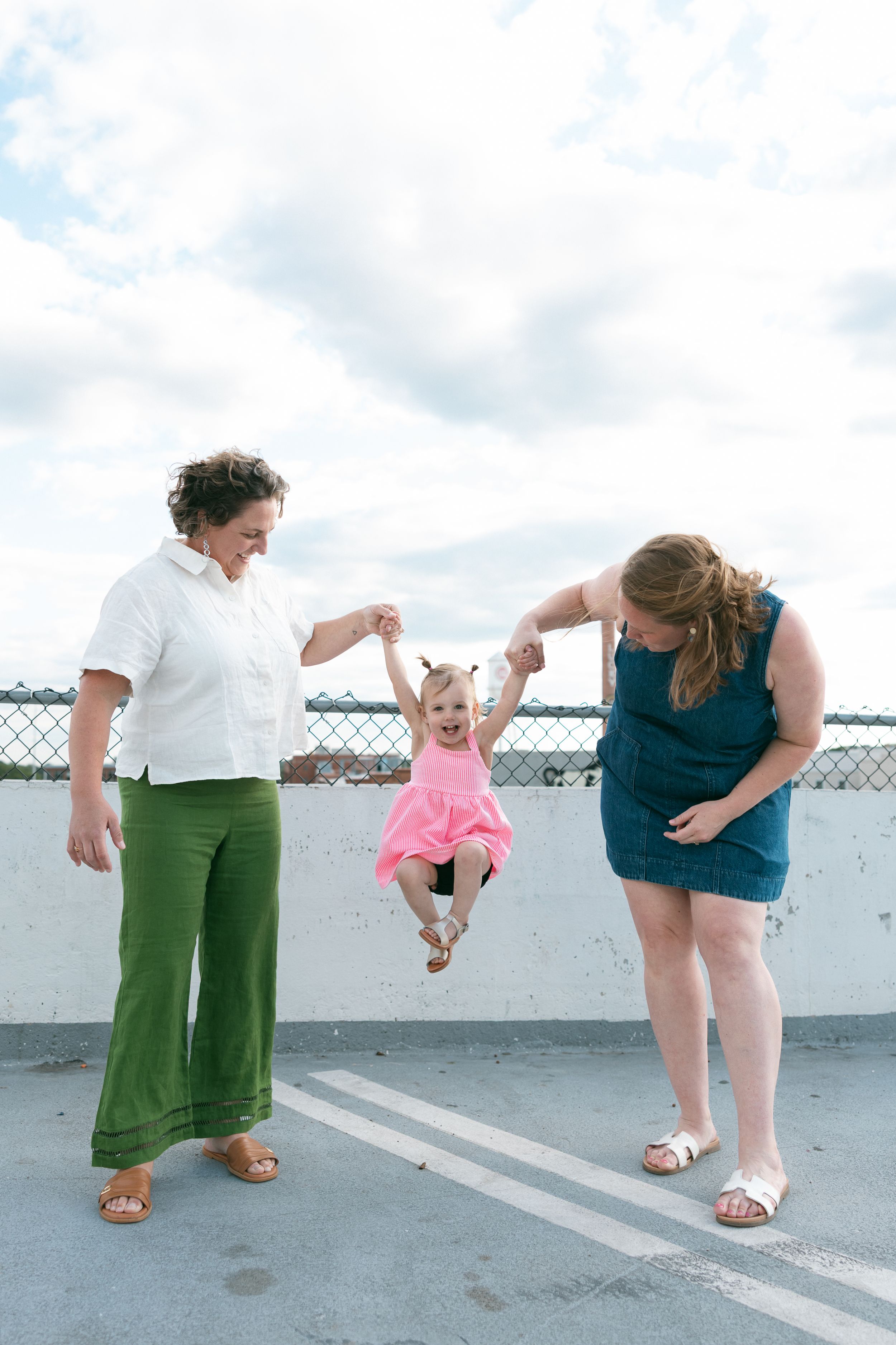 Two women swing a happy toddler girl in a pink dress between them on a rooftop under a cloudy sky.