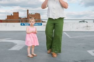 Toddler girl in pink dress stands beside adult in green pants on rooftop parking lot with city buildings behind.