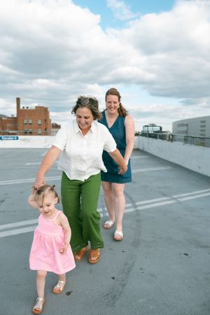 Two women and a toddler girl in a pink dress walk on a rooftop parking deck under a cloudy sky.