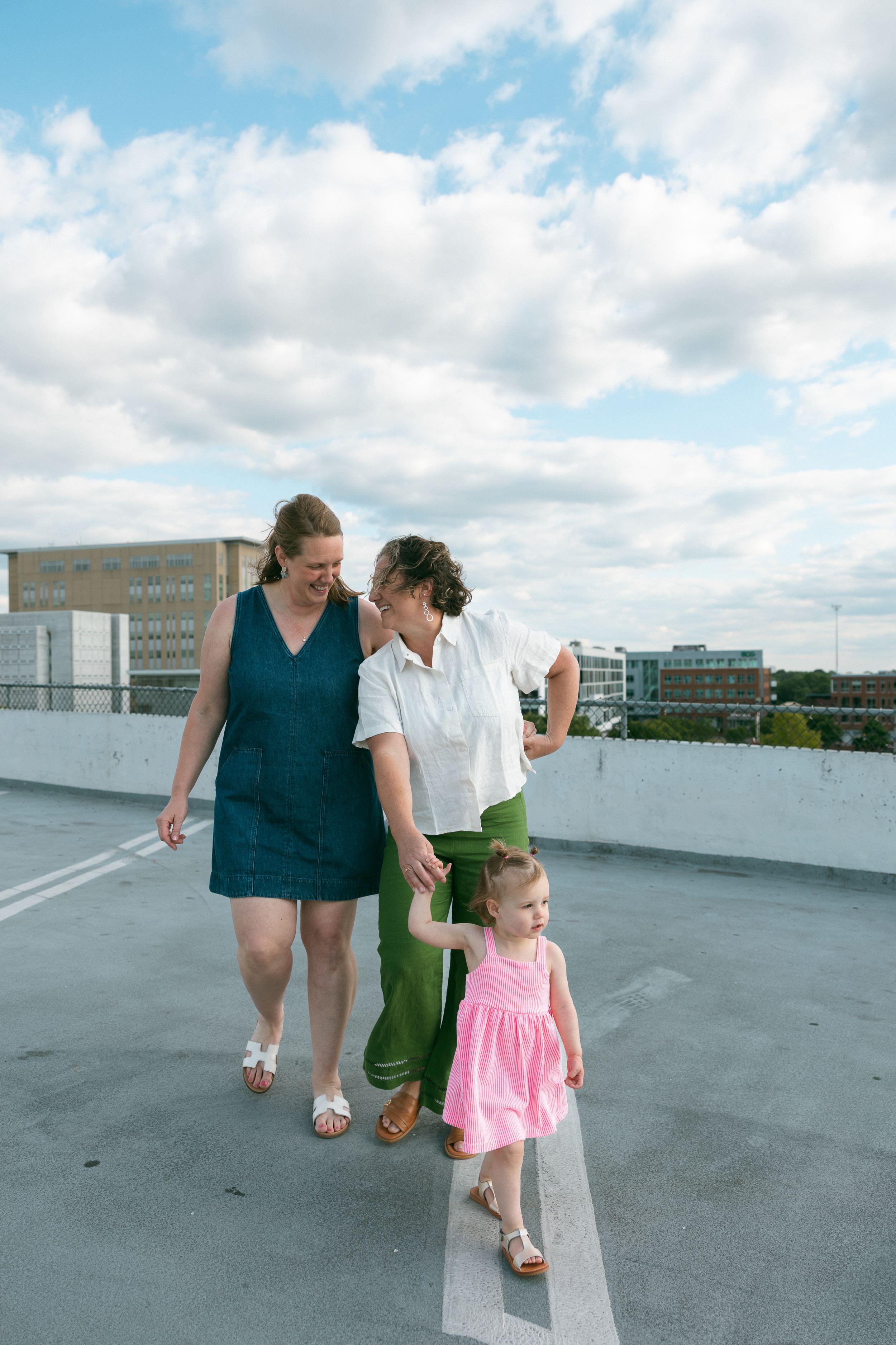 Two women and a young girl in a pink dress walk together on a rooftop parking lot under a partly cloudy sky.