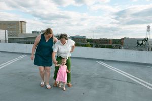 Two women and a toddler girl in pink dress hold hands on a rooftop parking lot with city skyline behind them.
