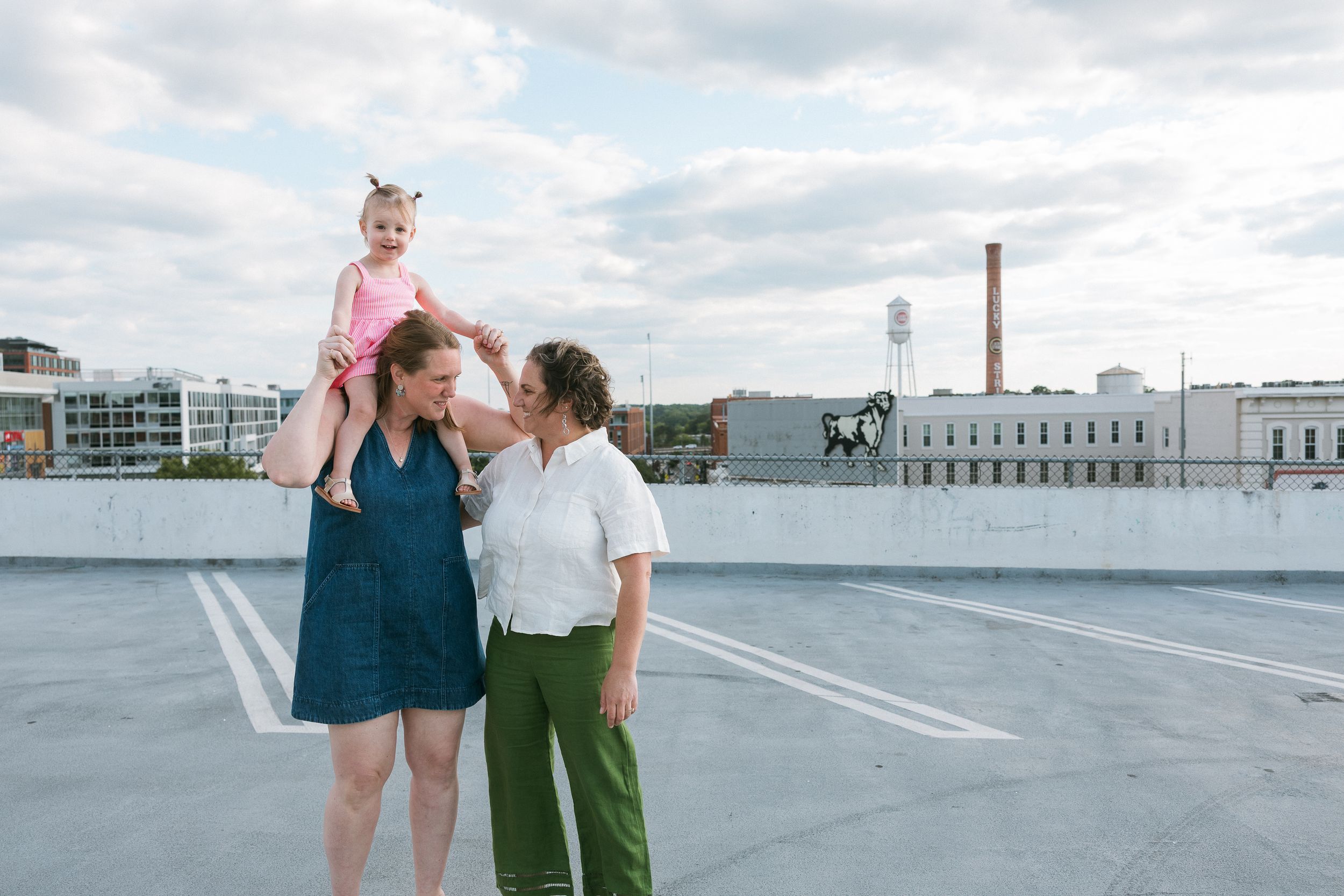 Two women and a toddler pose on a rooftop parking lot with a city skyline backdrop on a cloudy day.