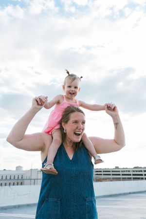 A joyful woman in denim overalls lifts a laughing toddler in pink onto her shoulders outdoors.