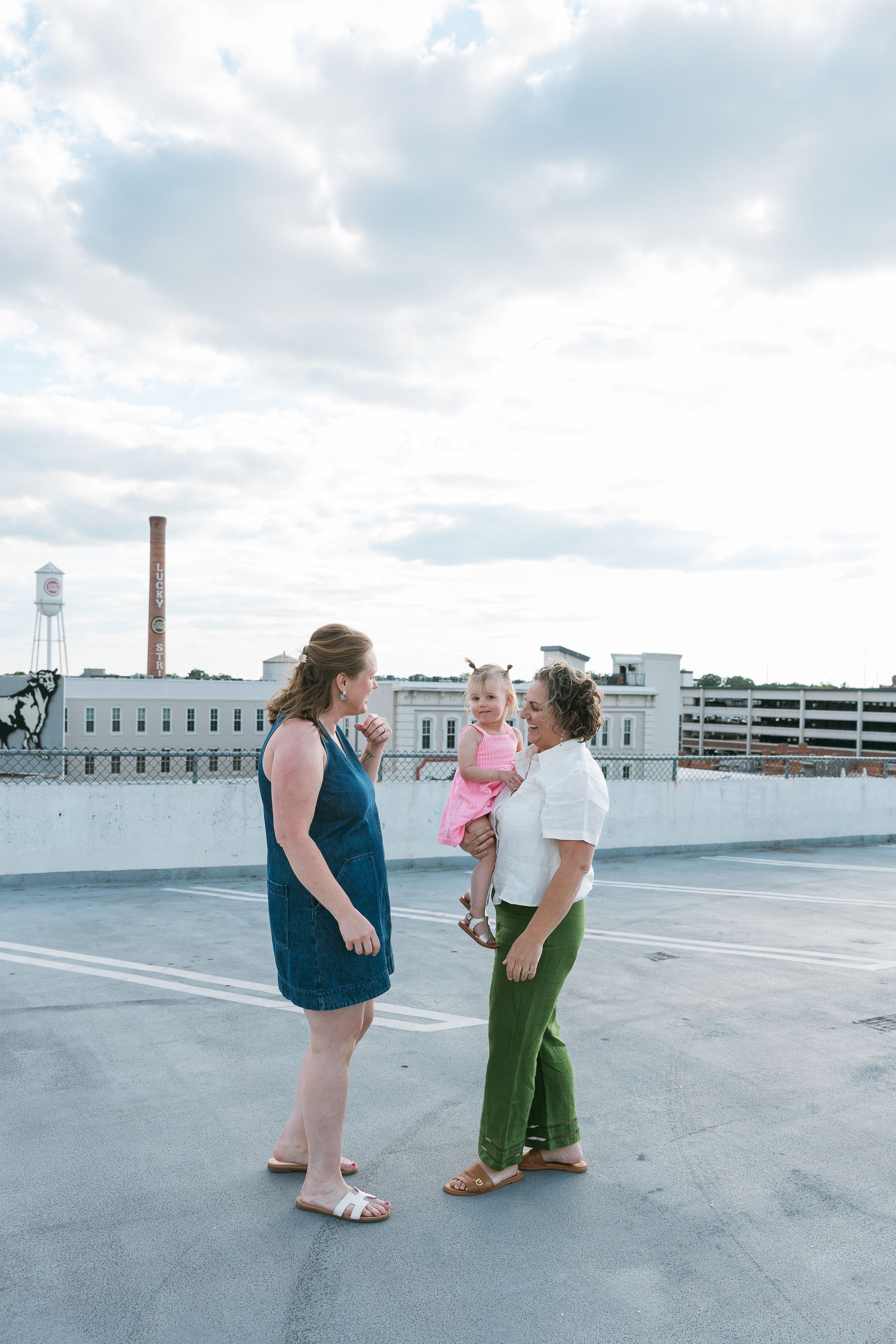 Two women and a toddler share a joyful moment on a rooftop parking deck with an urban skyline backdrop.