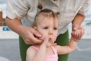Adult holding toddler girl in pink swimsuit outdoors, child sucking fingers while being supported by parent.