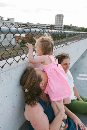 Two women sit against a chain-link fence on a bridge, holding a toddler girl in a pink dress exploring the fence.