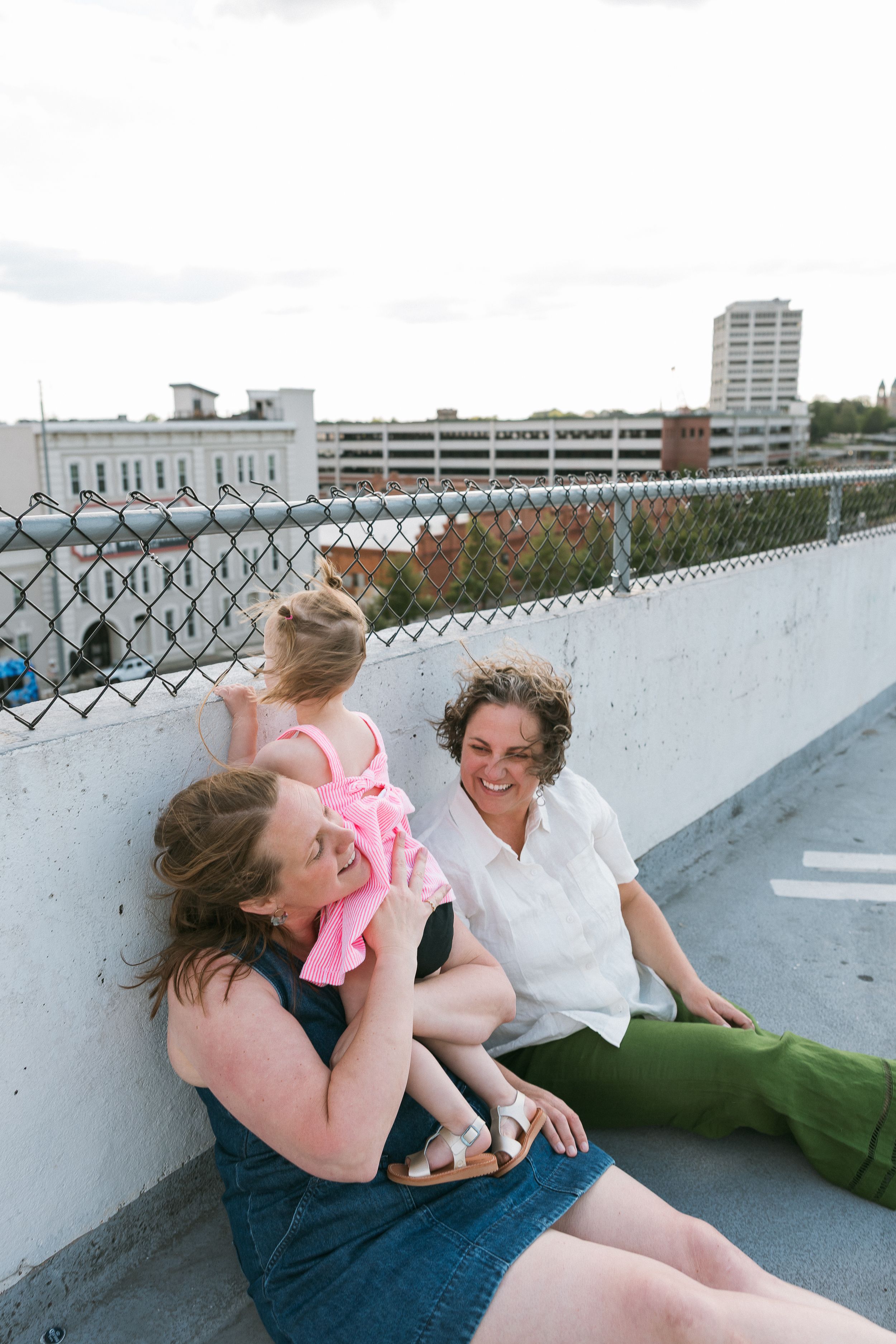 Two women and a toddler girl sitting together on a rooftop parking deck, laughing and enjoying a sunny day.