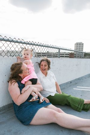 Two women and a toddler in pink sit together laughing on a rooftop parking structure.