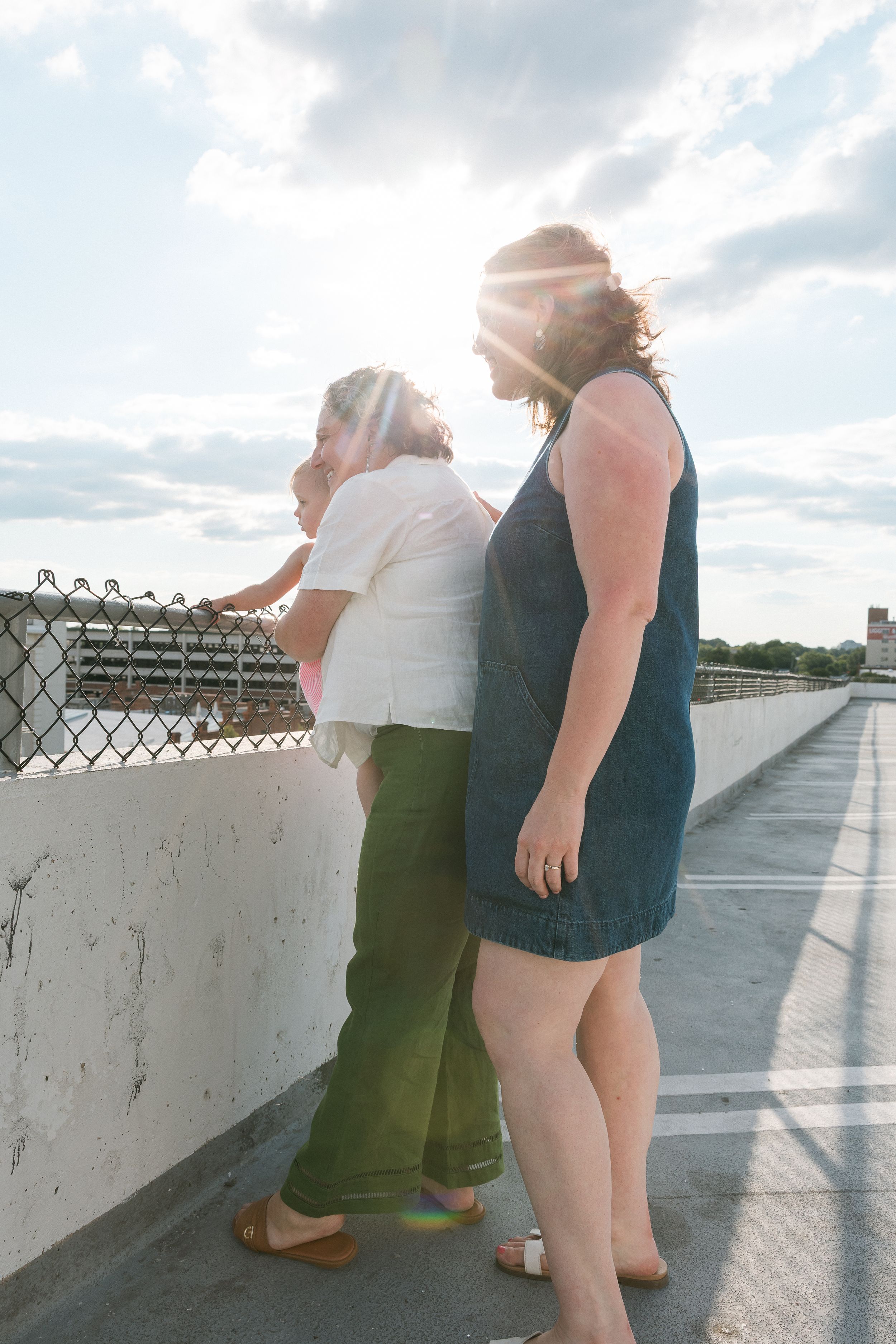 Two women standing on a bridge walkway in sunlight, one in white top with green pants, other in denim dress.