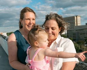 Two smiling women with a toddler girl in pink dress on a bridge, with city buildings in the background.