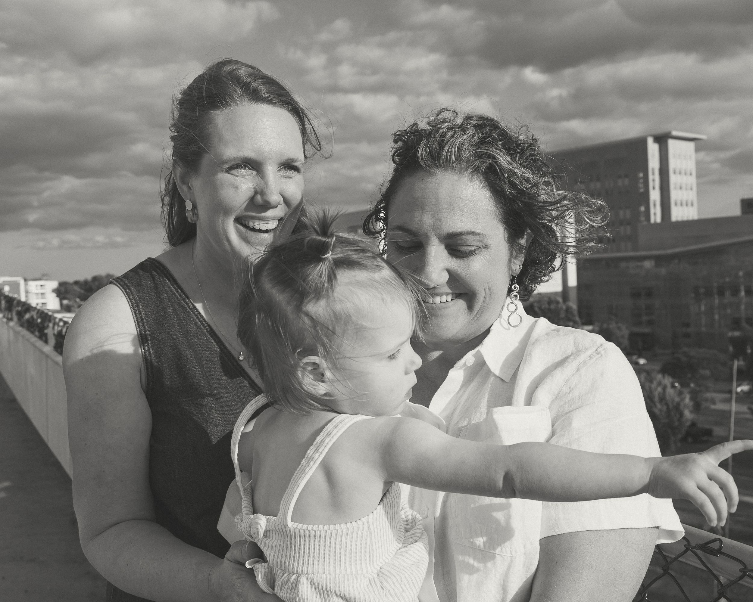 Black and white photo of two women smiling and holding a baby outdoors.