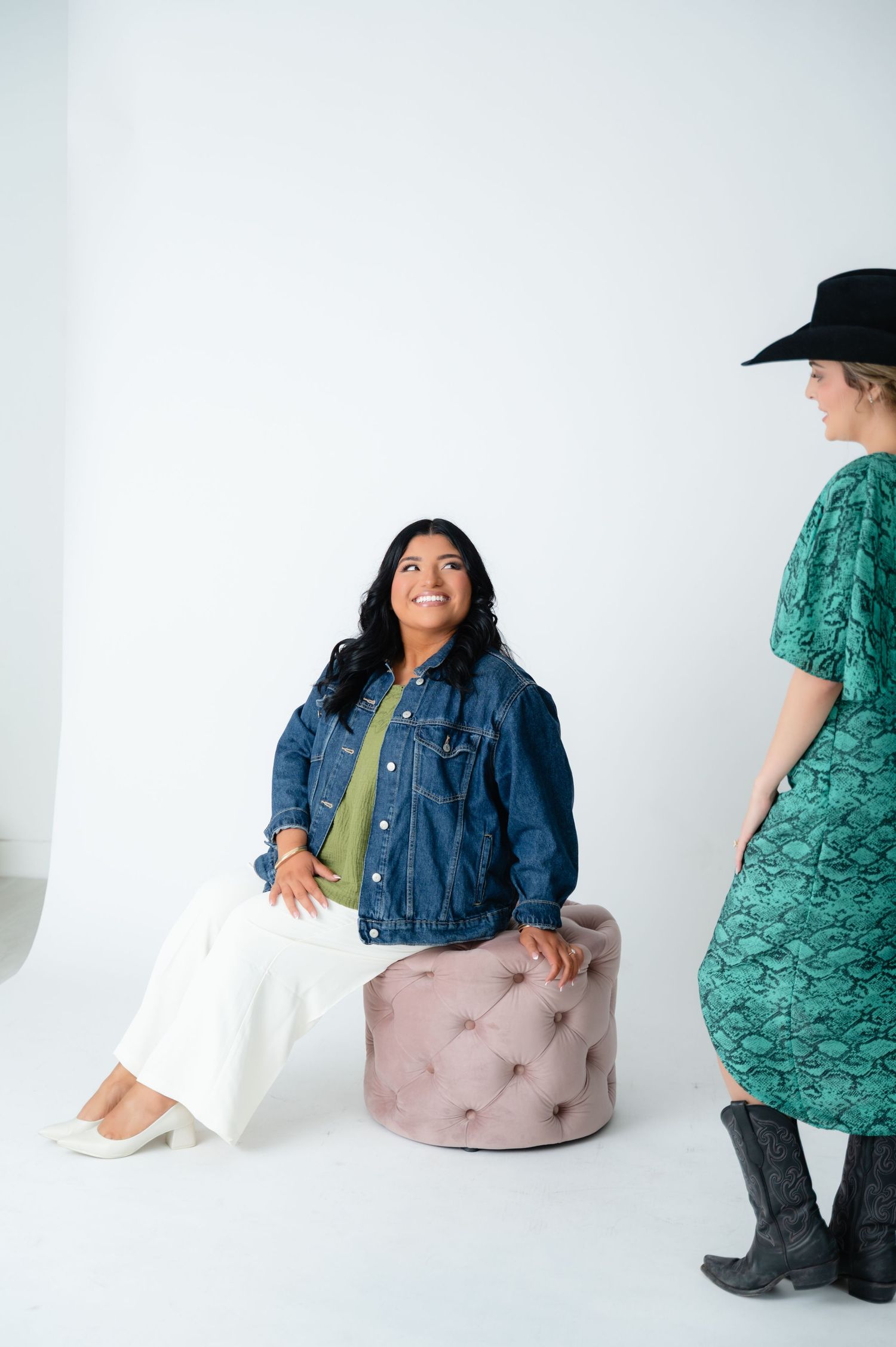 Woman in denim jacket sitting on tufted ottoman smiling at person in cowboy hat.