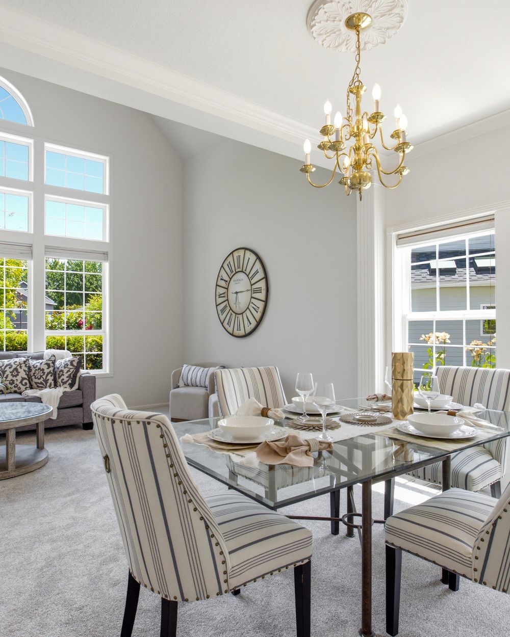 Elegant dining room with glass table, striped chairs, gold chandelier, and large windows in a bright gray interior.