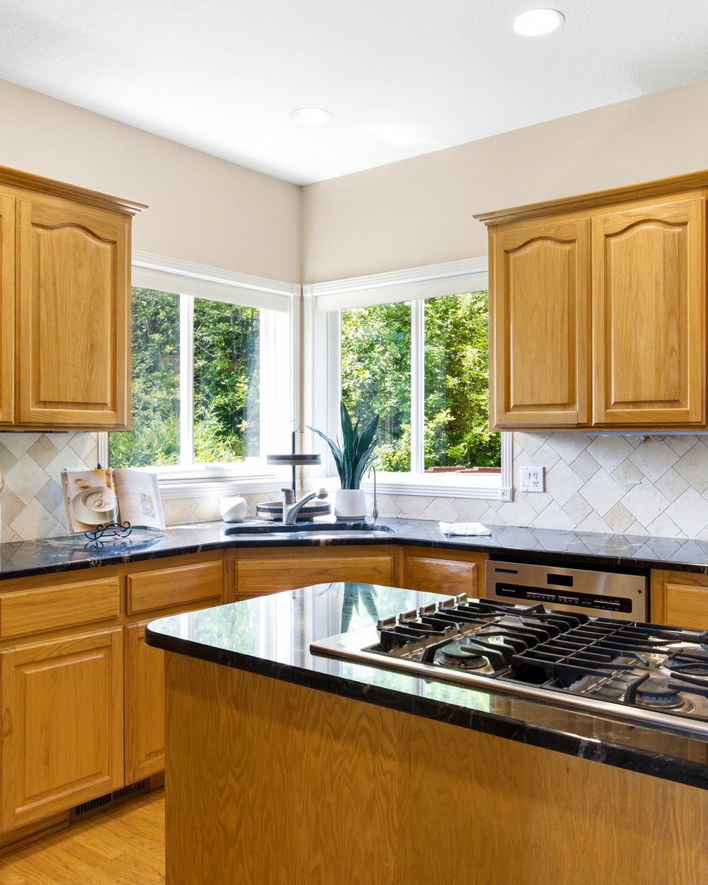 Bright kitchen with oak cabinets, black granite countertops, gas stove, tile backsplash, and large windows overlooking greenery.