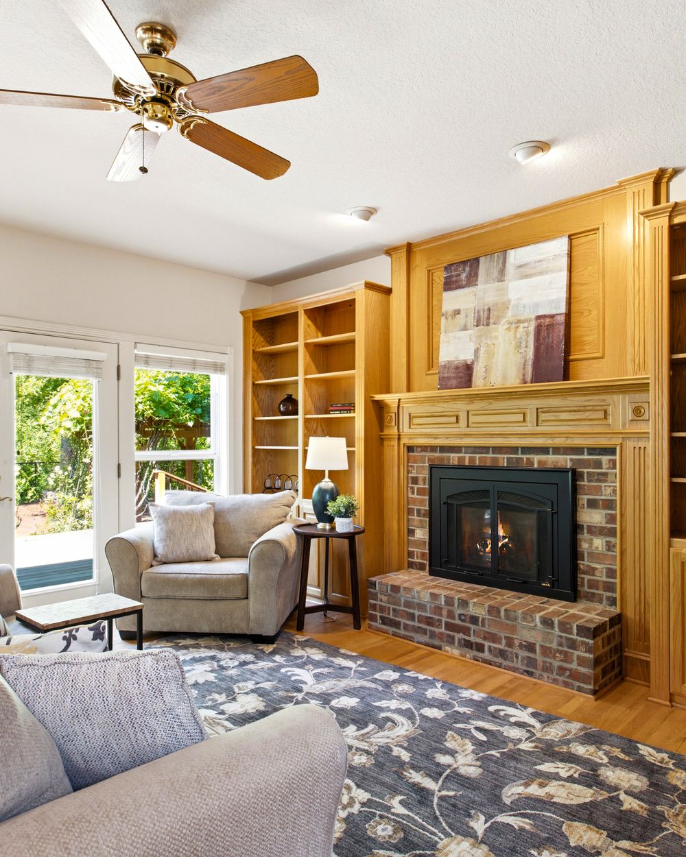 Cozy living room featuring a brick fireplace, built-in wood shelving, ceiling fan, and hardwood floors with natural light.