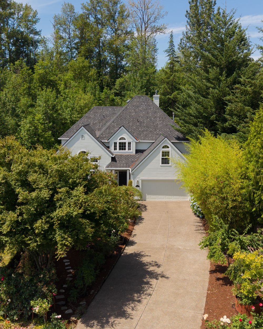 Aerial view of a blue-gray two-story home with a gray roof, surrounded by lush green trees and a concrete driveway.