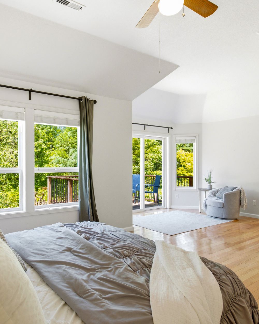 Bright master bedroom with hardwood floors, large windows, ceiling fan, and French doors opening to a deck with lush green views.