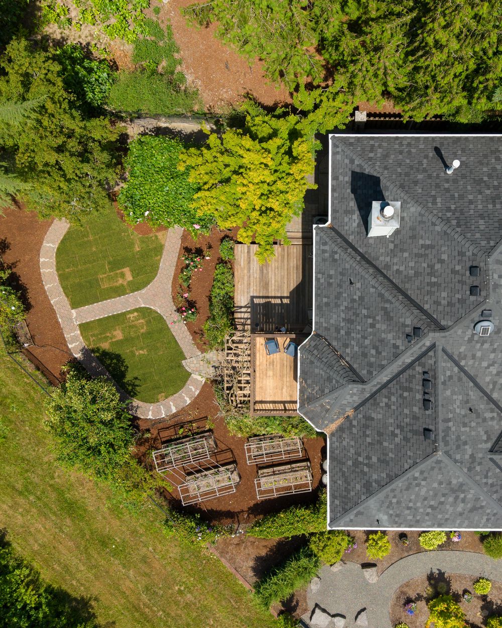 Aerial view of a residential property featuring a gray shingle roof, wooden deck, and landscaped backyard with curved pathways.