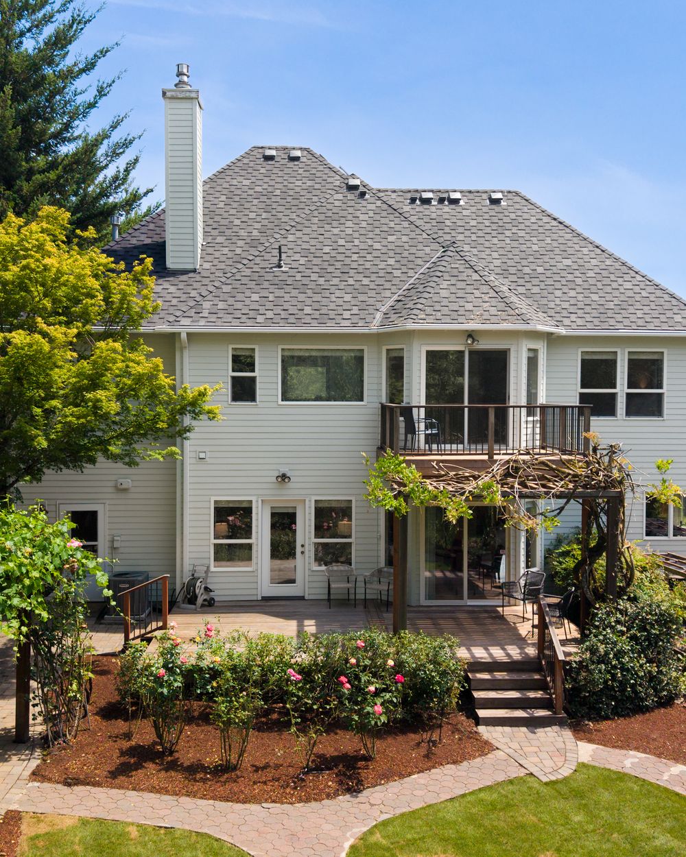 Two-story white home with gray roof, wraparound deck, pergola, and lush landscaped garden with rose bushes.