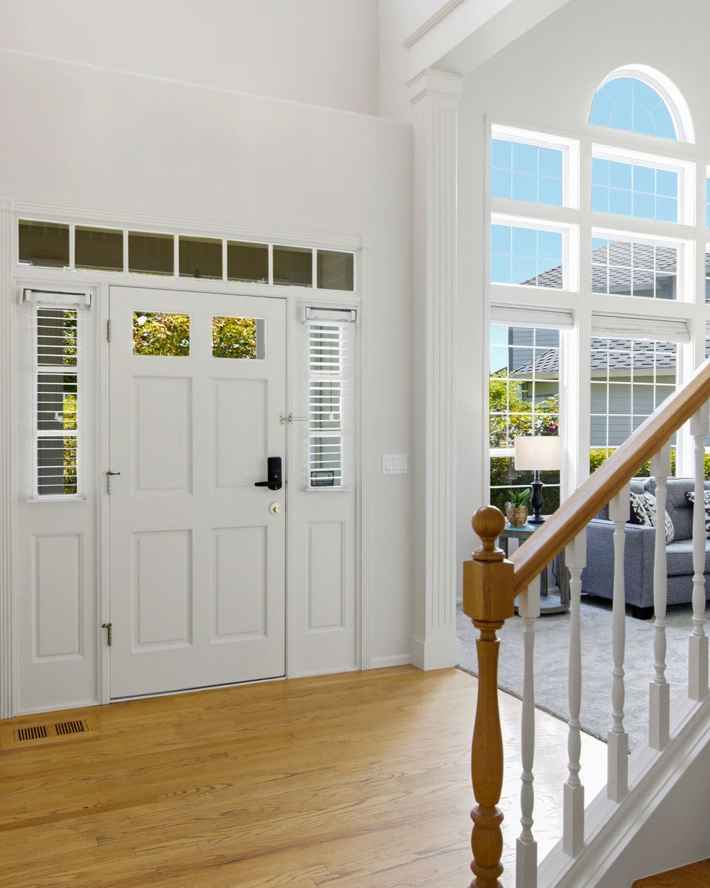 Bright home entryway with white front door, hardwood floors, arched window, and staircase with wood railing.