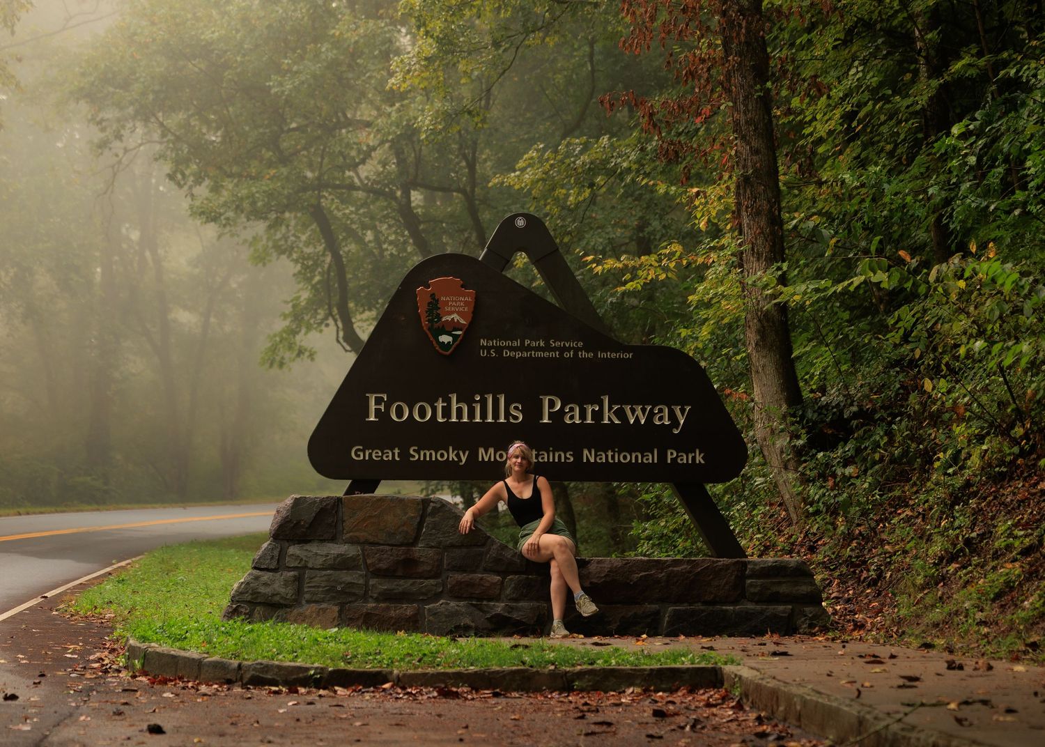 Woman posing at Foothills Parkway sign, Great Smoky Mountains National Park, surrounded by misty foggy trees.