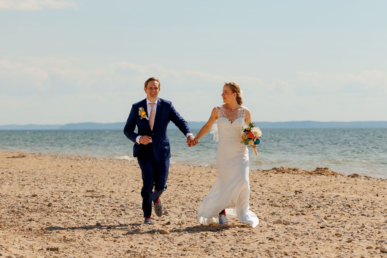 Bride and groom holding hands running on a sandy beach on their wedding day with ocean in background.