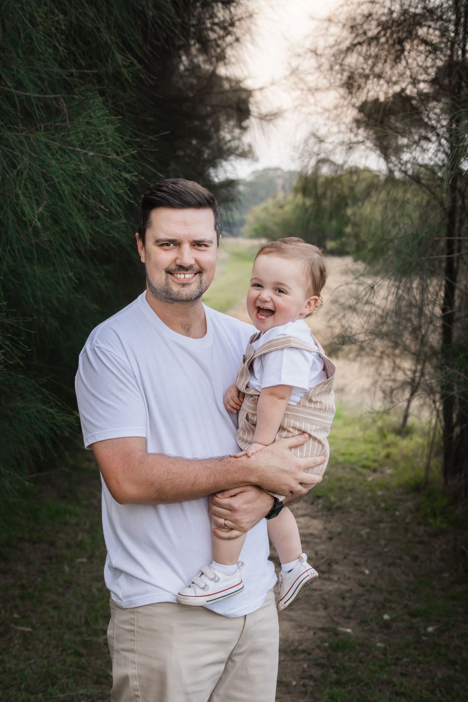 A smiling man in a white t-shirt holds a laughing baby boy outdoors at Rouse Hill Regional Park among green trees archway.