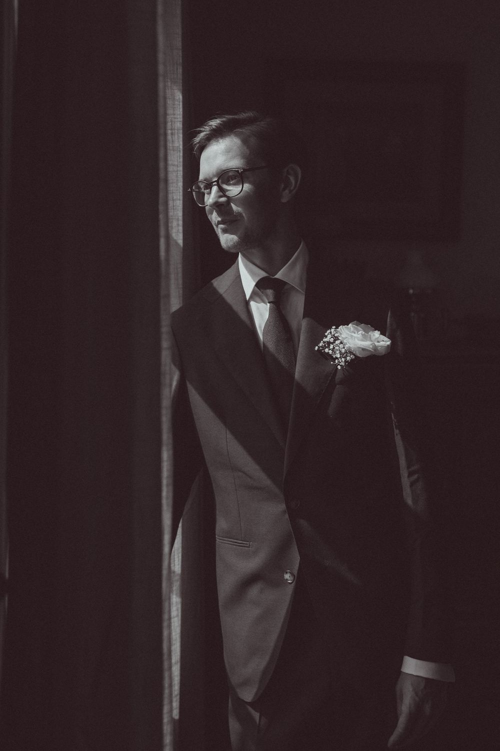 Black and white portrait of a smiling groom in a suit with boutonniere, standing by a window in soft light.