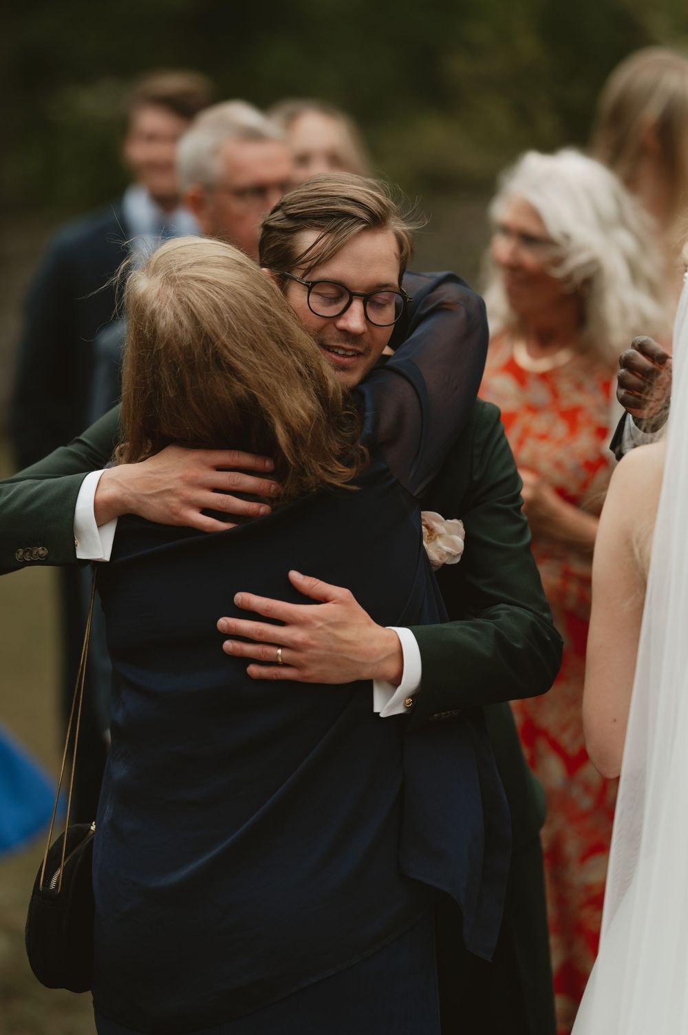 A young man wearing glasses shares a warm embrace with a guest at an outdoor wedding celebration.