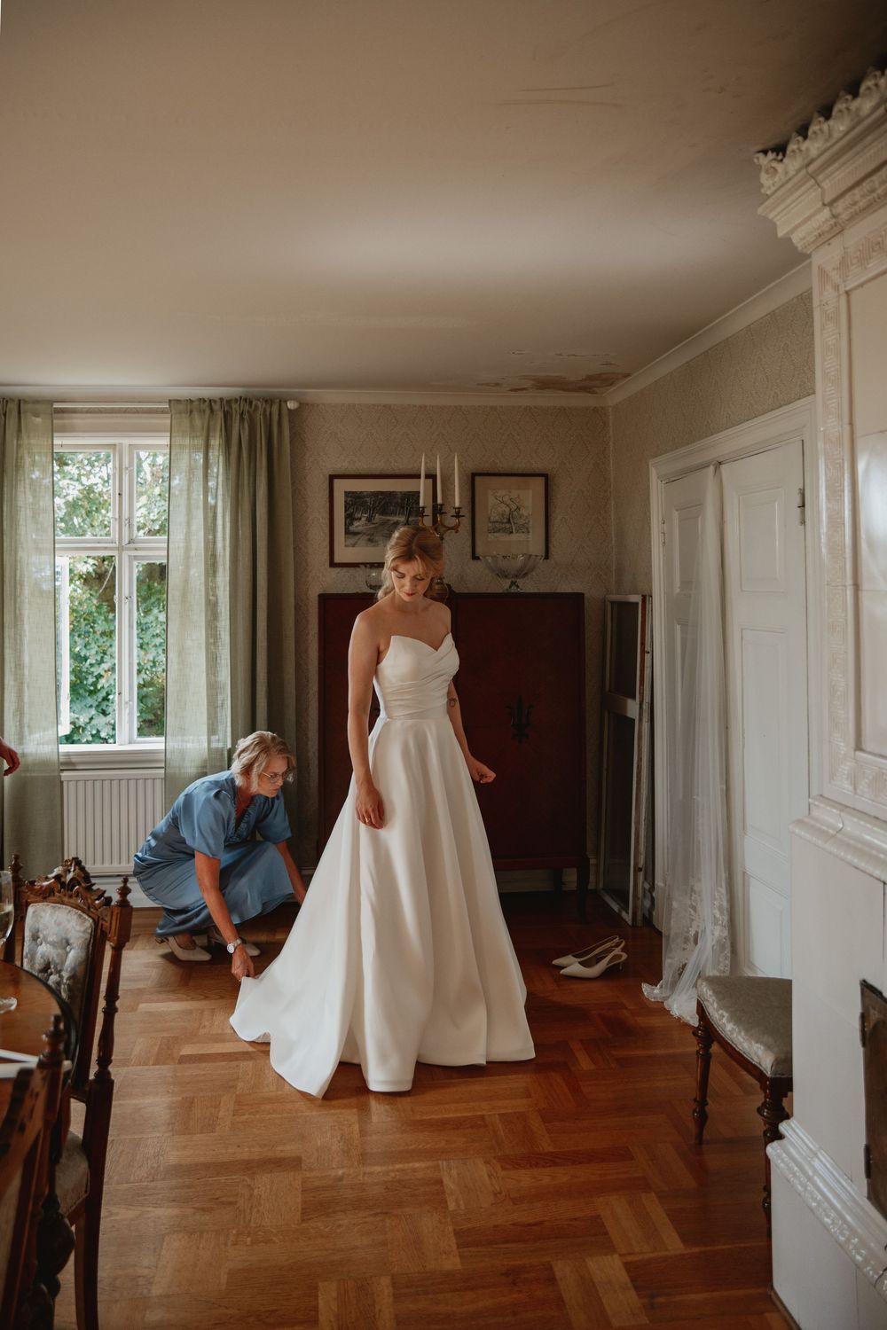 Bride in elegant white A-line wedding gown being helped with shoes in a classic wood-floored room.