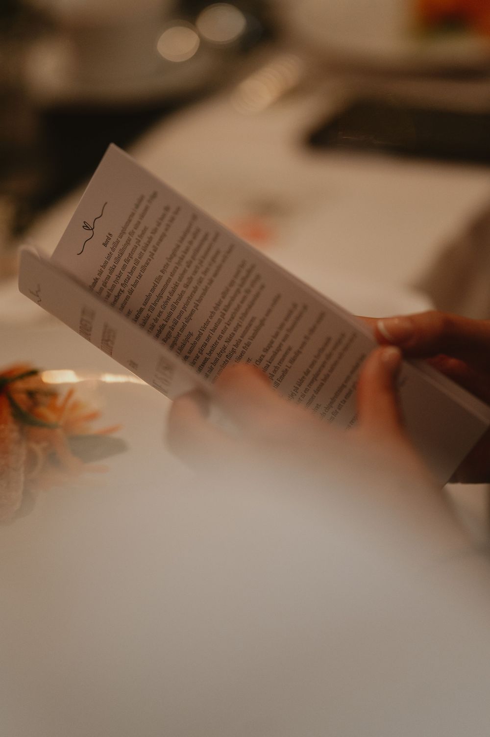 Person holding a printed wedding menu card at a reception table with floral decorations in soft warm lighting.