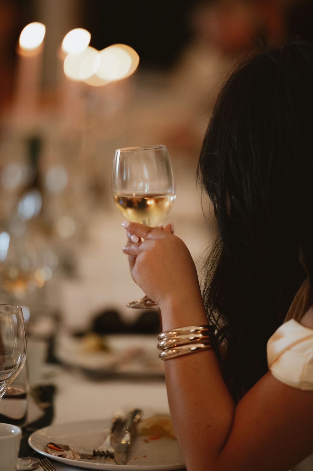Woman holding a glass of white wine at an elegant candlelit dinner party, wearing a gold bracelet.