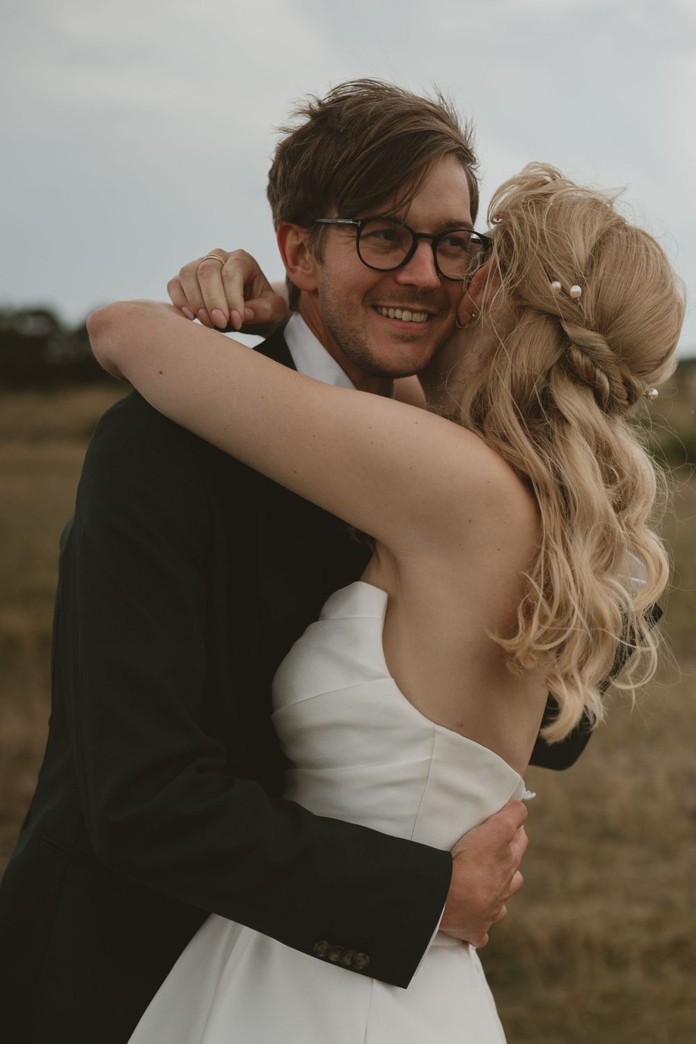A smiling groom in glasses embraces his bride in a white dress outdoors on their wedding day.